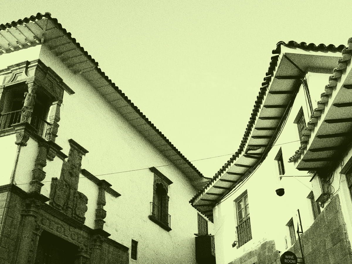 Looking up at the tiled eaves of two colonial houses in Cusco, Peru, one with a carved stone balcony and doorway.